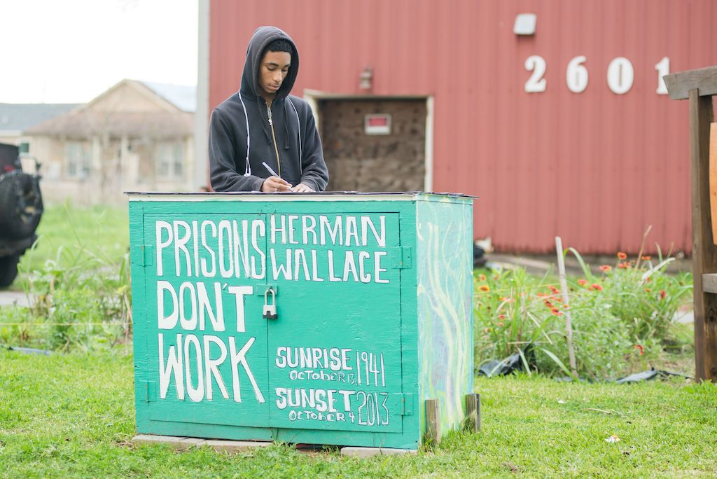 man wearing a black hoodie is standing behind a teal wooden box that says "prisons don't work' in white letters.