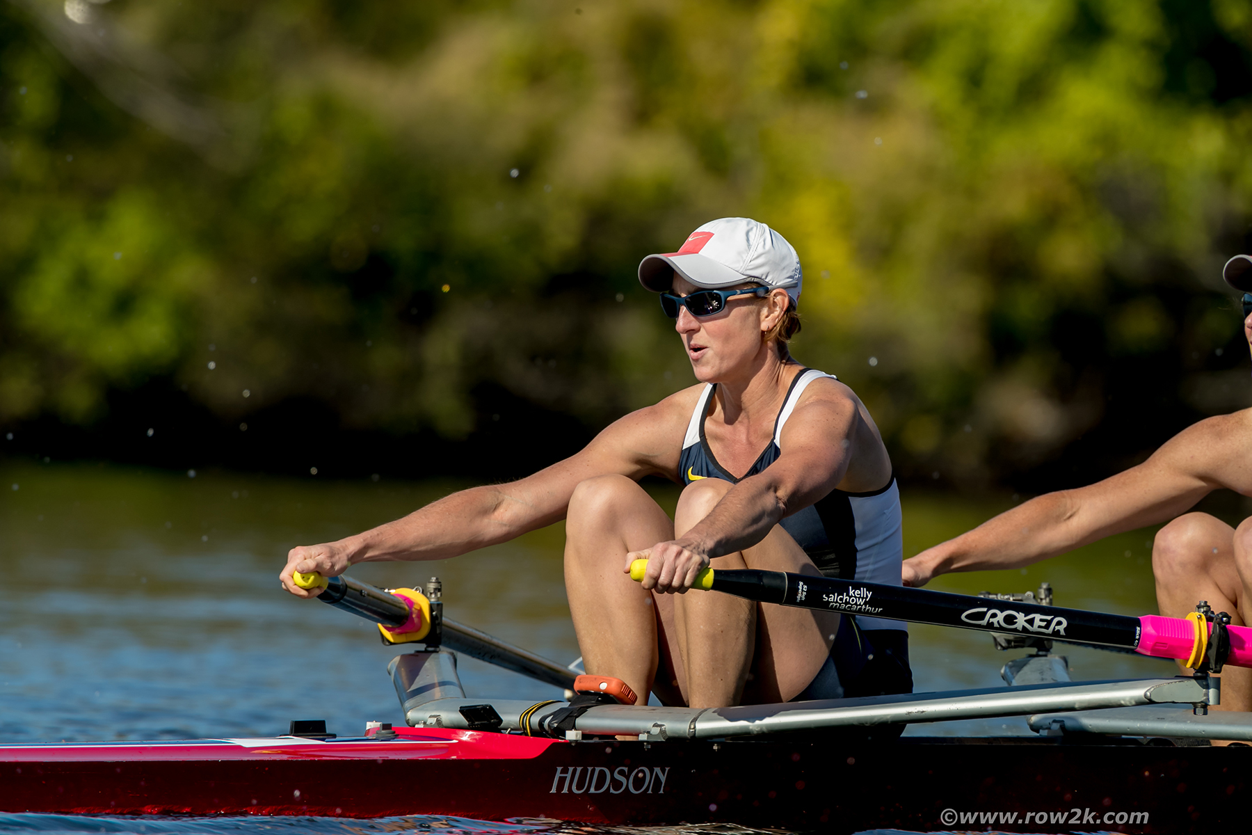 A woman with sunglasses and a baseball cap wearing athletic clothes rowing in a rowboat on a lake.