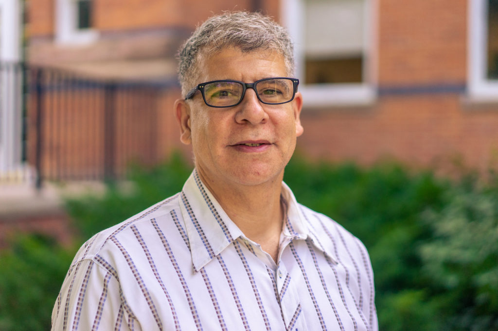 A portrait of a smiling man wearing glasses and a button-down shirt outdoors.