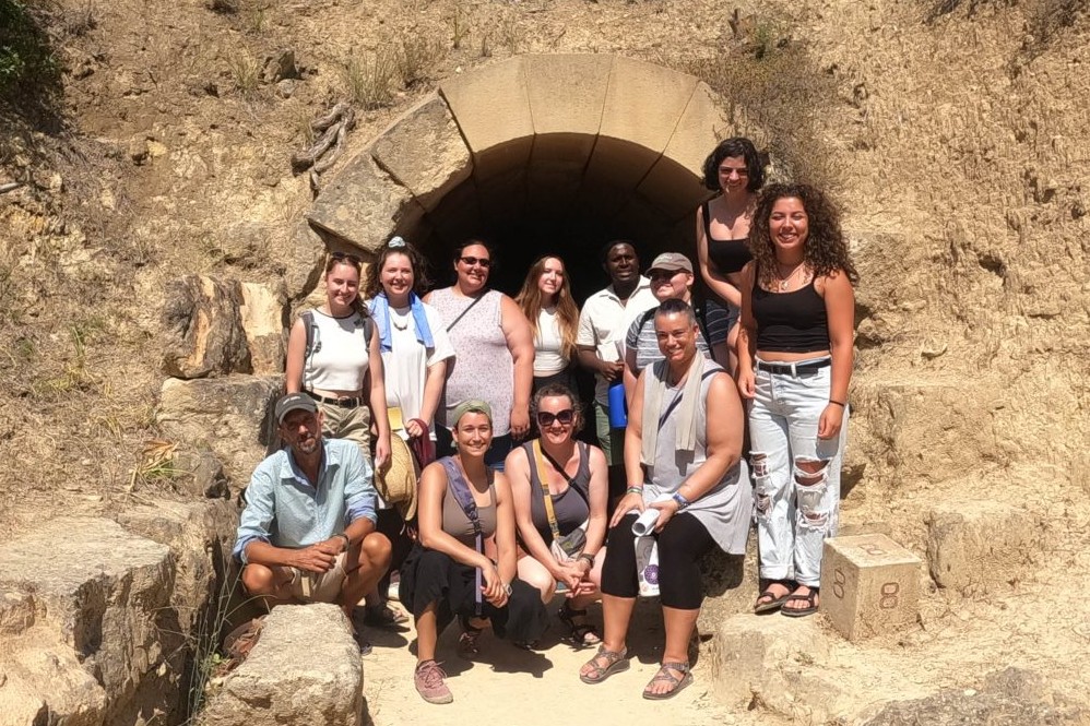 Ten people gathered outdoors in front of cave opening, smiling at camera.