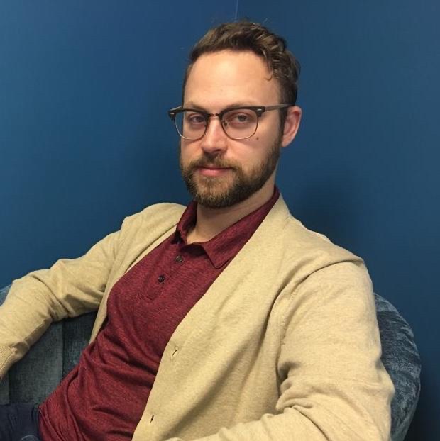 A picture of a man in tan cardigan and a red polo in front of a blue background.