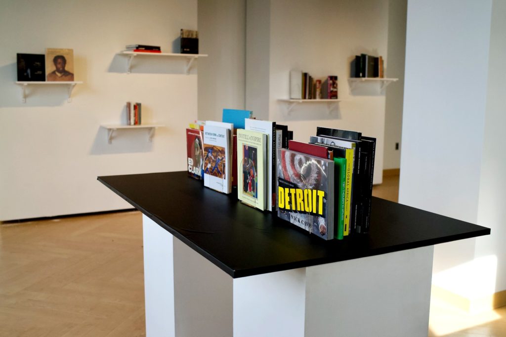 Table with black colored top with several books on top. In the background are shelves hanging on the walls wthe a few books on each shelf. 