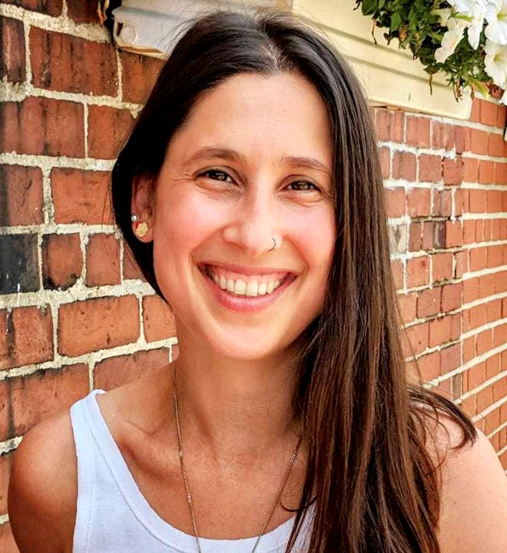 A picture of a woman in a white tank-top with long brown hair smiling and posed in front of a brick wall.