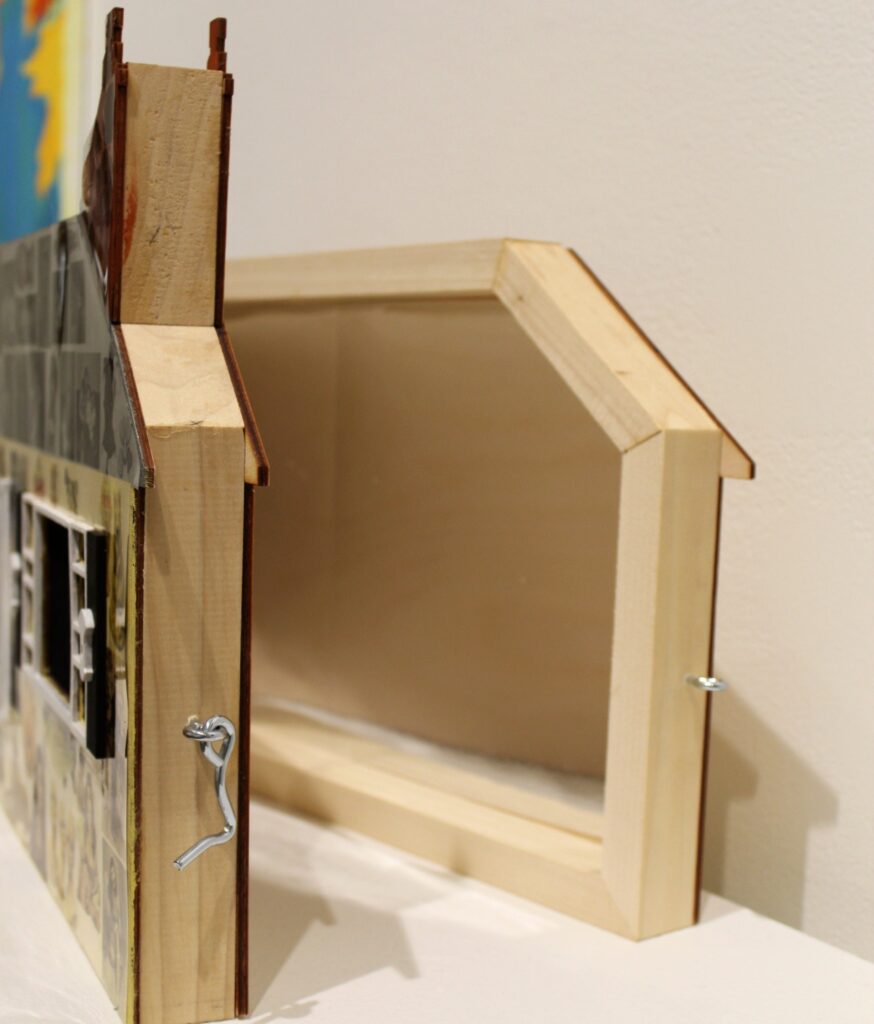 A side view of a house-shaped sculpture showing its wooden structure and metal hinges.