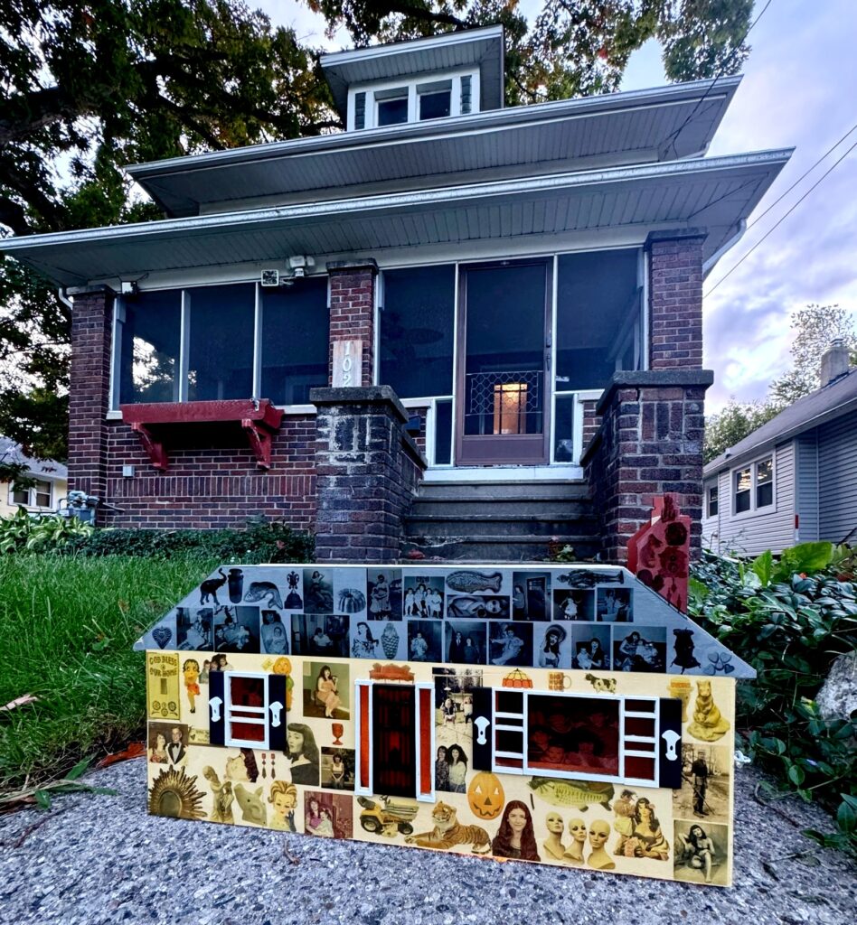 A picture of a mixed-media house sculpture placed on a concrete path in front of a brick house. 
