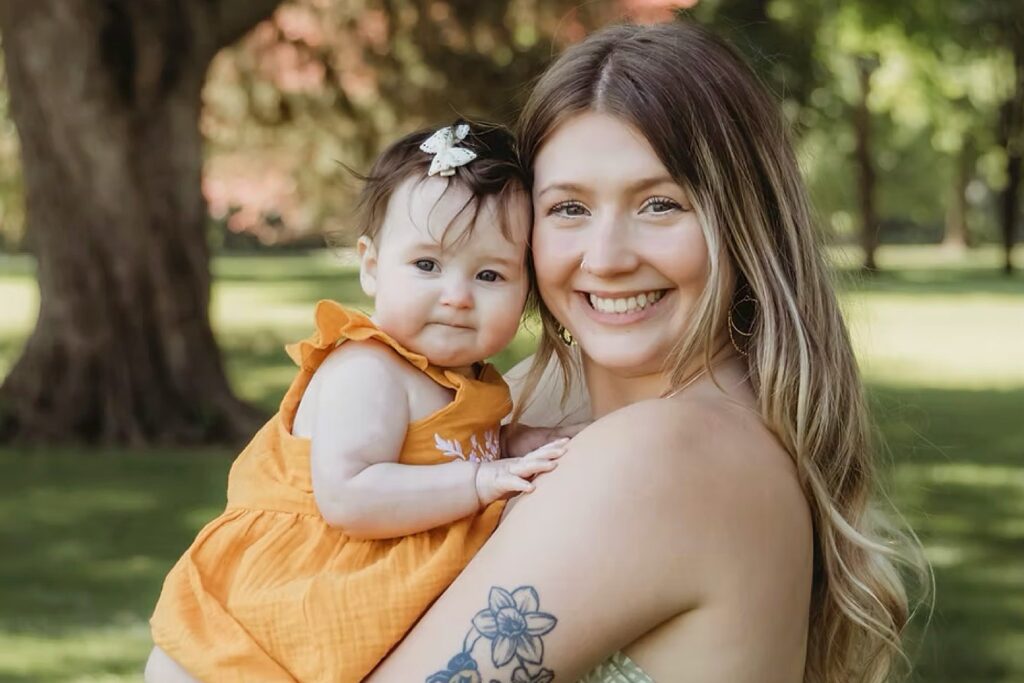 Woman with long blonde hair and smiling holding a baby girl with short brown hair and a bow. 