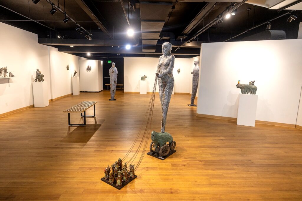 A wide-angle view of a gallery featuring large, textured ceramic sculptures on pedestals and walls. The centerpiece is a tall figure pulling a wheeled creature that is connected to a cluster of small, colorful figurines on the floor.