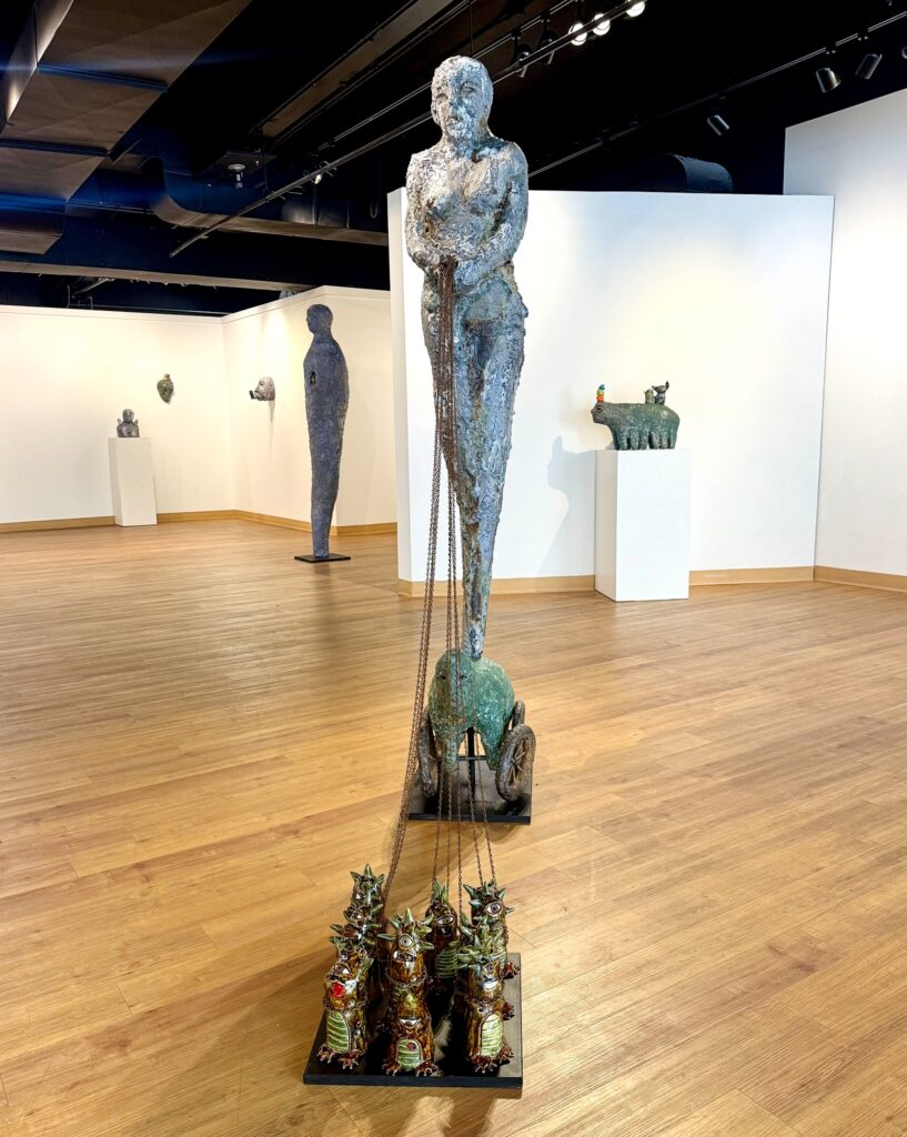 A tall human figure sculpture holding chains that are connected to whimsical, glossy ceramic creatures arranged on a black base. Each has a horned, reptilian form with chains draped across their bodies. 