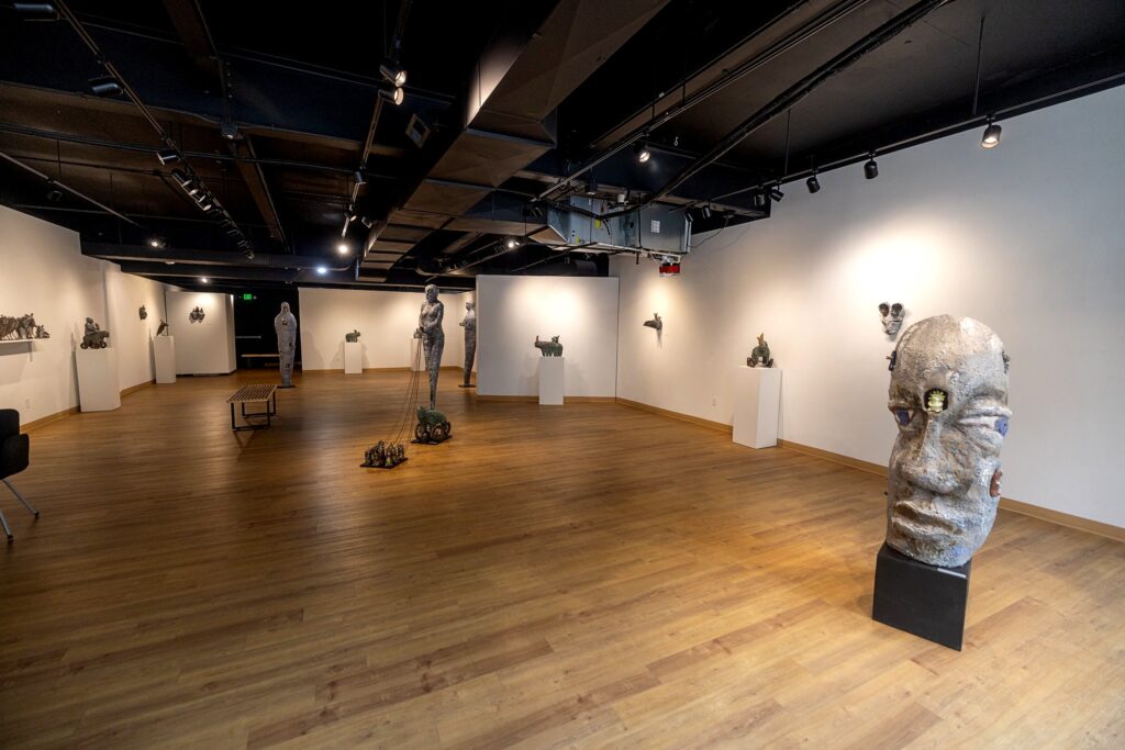 A picture of an art gallery with wooden floors and white walls displaying a variety of humanoid and animal-like  ceramic sculptures.