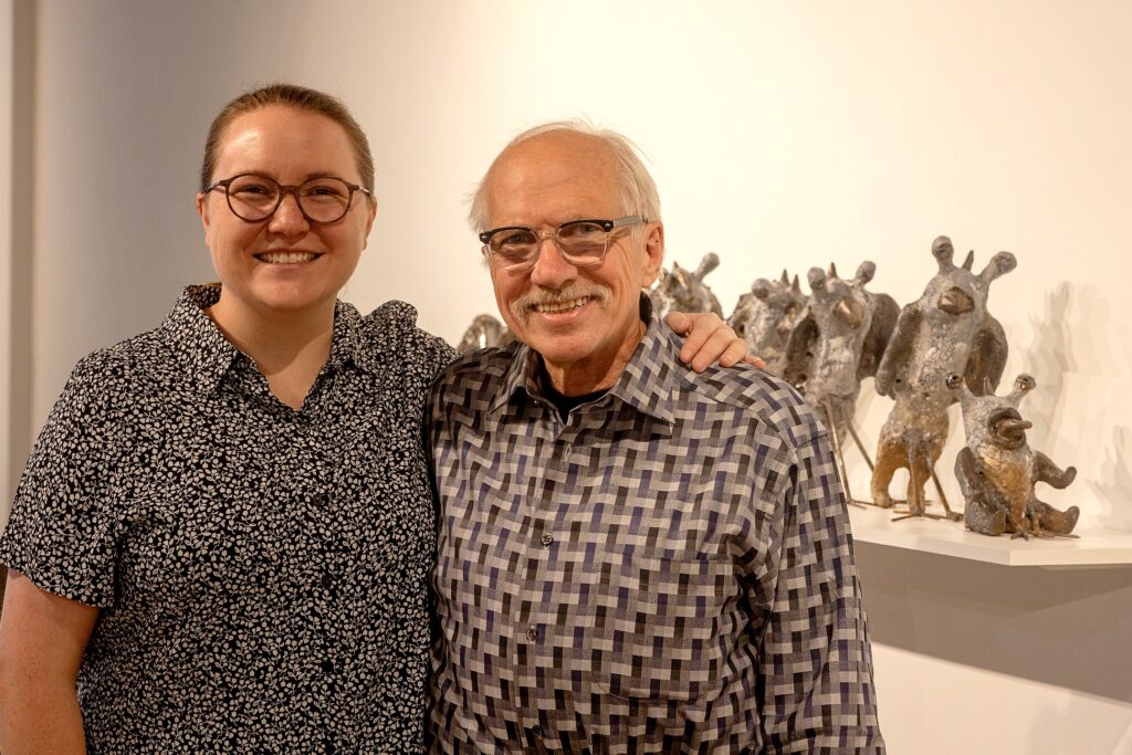 Two people stand smiling in a gallery. The person on the left has their arm around the person on the right. To the right is a shelf full of small, creature-like figures..