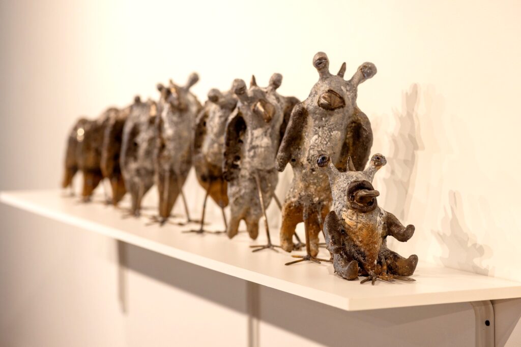 A row of whimsical bird-like sculptures made of textured, metallic-looking material displayed on a white shelf. Each figure has distinct facial expressions and small, upright protrusions resembling antennae or ears.