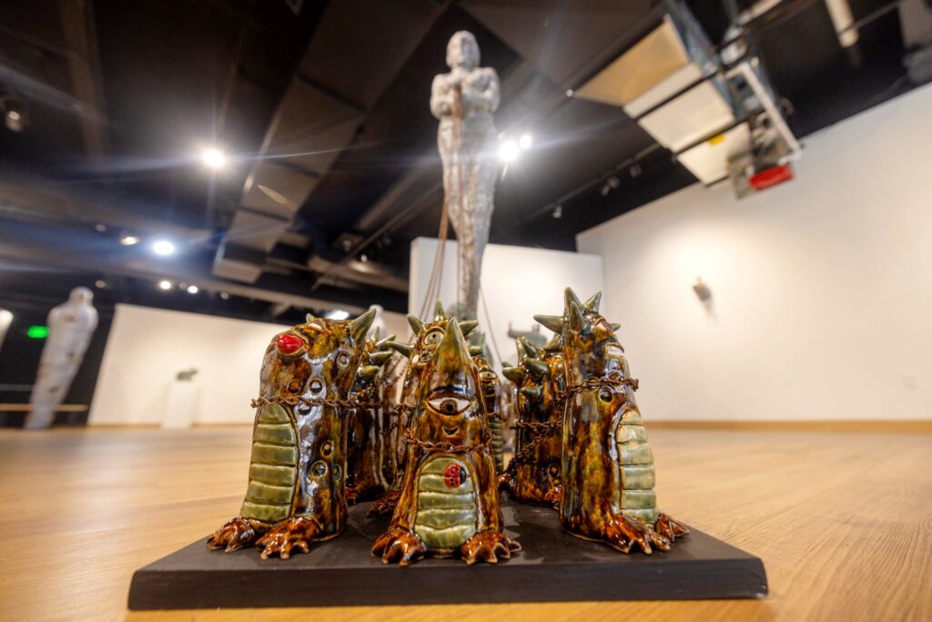 Low-angle view of a cluster of whimsical, glossy ceramic creatures arranged on a black base. Each has a horned, reptilian form with chains draped across their bodies. The background shows a tall figure sculpture holding the chains.