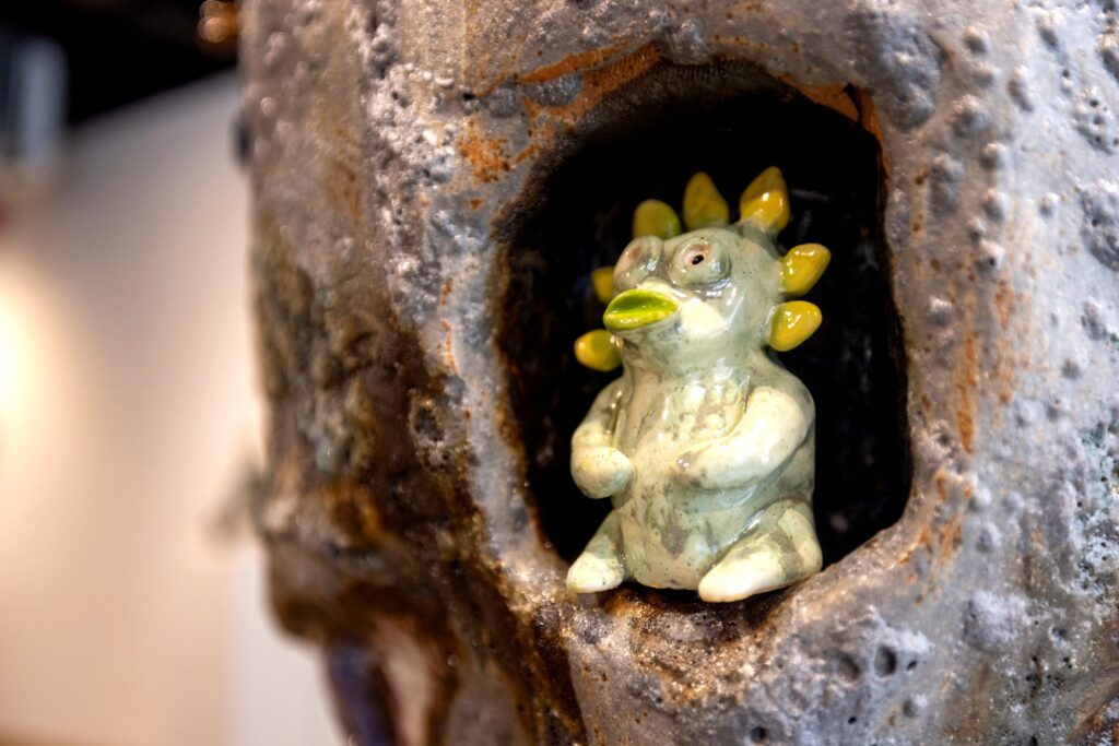 A light green, glossy ceramic creature with yellow spike-like hair and wide eyes sits inside a hollowed-out section of a larger sculpture.