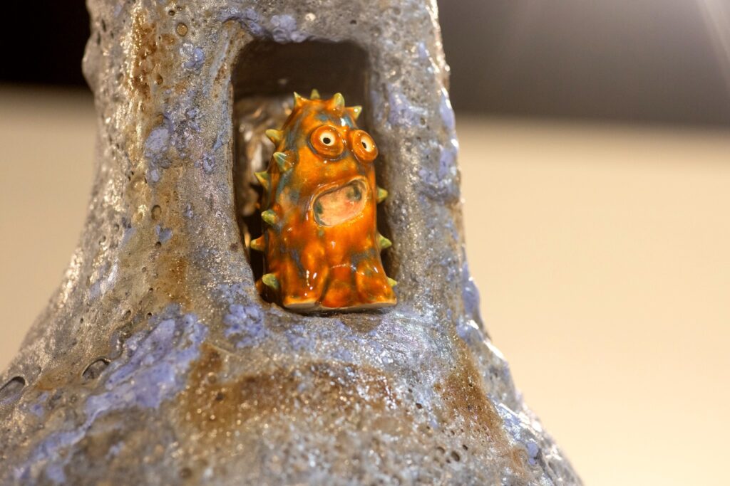 A close-up of a bright orange, spiky ceramic creature with large eyes and an open mouth. It is nestled into a alcove of a tall, gray and blue textured sculpture.