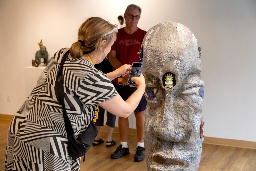 A woman taking a photo with her cell phone of a large ceramic sculpture of a human head that is textured with exaggerated facial features and small, colorful creatures embedded in its surface.