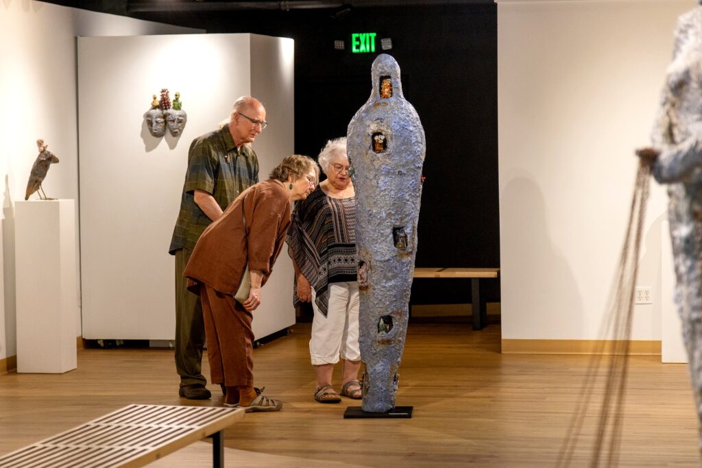 Three people, one man and two women, looking at a tall, cocoon-shaped sculpture with embedded miniature figures in caves along its surface. The piece stands upright on a black pedestal in the center of a gallery room.
