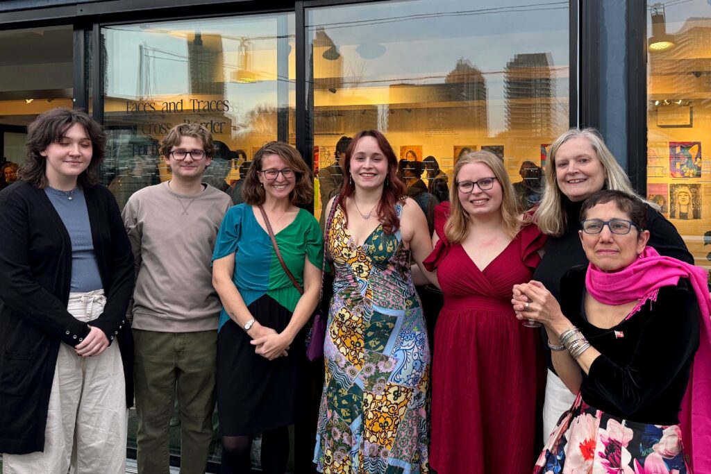 Seven people stand smiling outside a gallery with the exhibition title “Faces and Traces: A Cross-Border Collaboration” visible on the window behind them. Reflections of city buildings appear in the glass.
