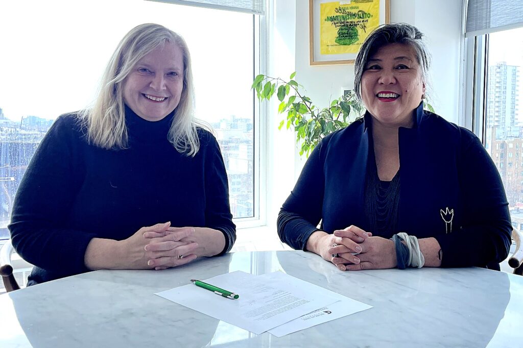 Two women sit at a round marble table in front of large windows with a city view, smiling toward the camera. A signed document and green pen rest on the table between them.