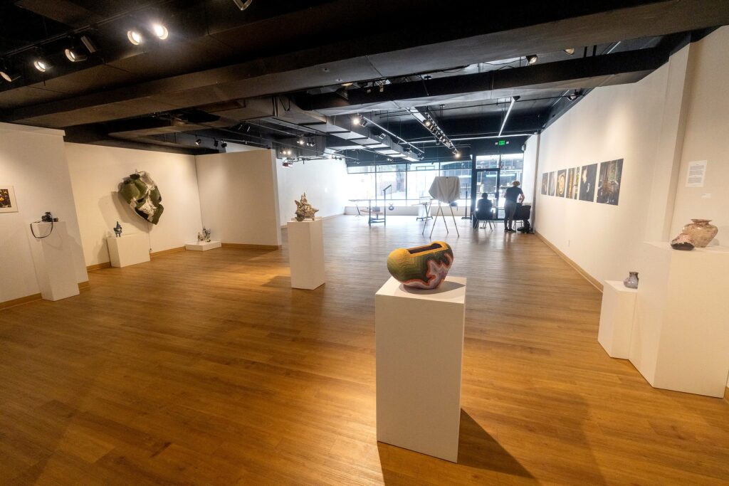 A wide view of the “Nexus” exhibition gallery showing ceramic sculptures, photographs, and mixed-media artworks displayed on white pedestals and walls under track lighting.