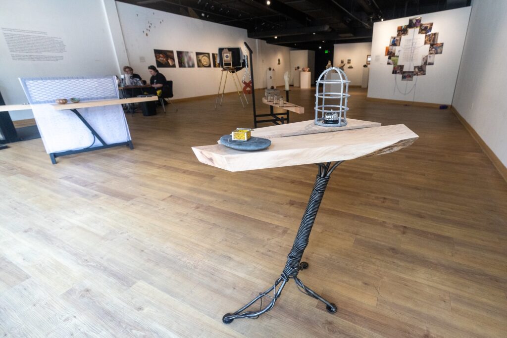A sculptural installation sits on a tall wooden pedestal with a metal base. On top, a wire cage structure and small objects, including a yellow box, rest on the surface. In the distance, visitors sit at a table while additional artworks and photographs line the gallery walls.