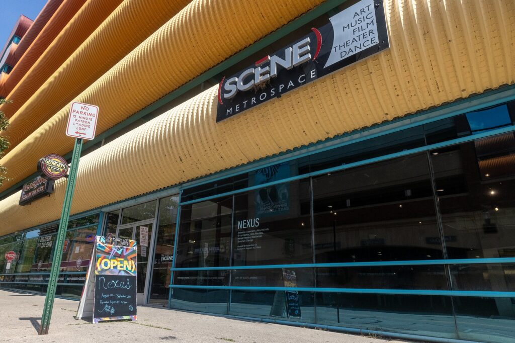 Exterior of the (SCENE) Metrospace gallery in East Lansing. A sidewalk sign reads “Nexus, August 17–September 21” with the reception date and hours. The gallery’s name is displayed in large letters above the entrance, alongside colorful building panels.