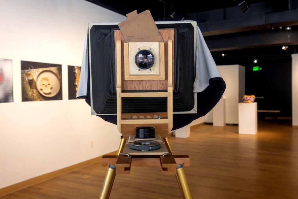 A large-format wooden camera with a bellows stands on a tripod inside the gallery. The camera faces the viewer, with framed photographs displayed on the wall behind it.