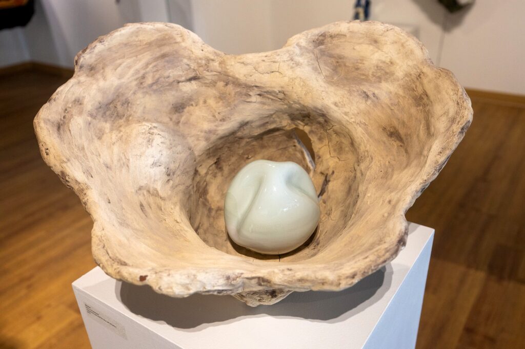 A sculptural piece resembling a large, organic wooden oyster sits on a pedestal. Inside the hollowed form rests a smooth, pale green ceramic sphere with gentle folds.