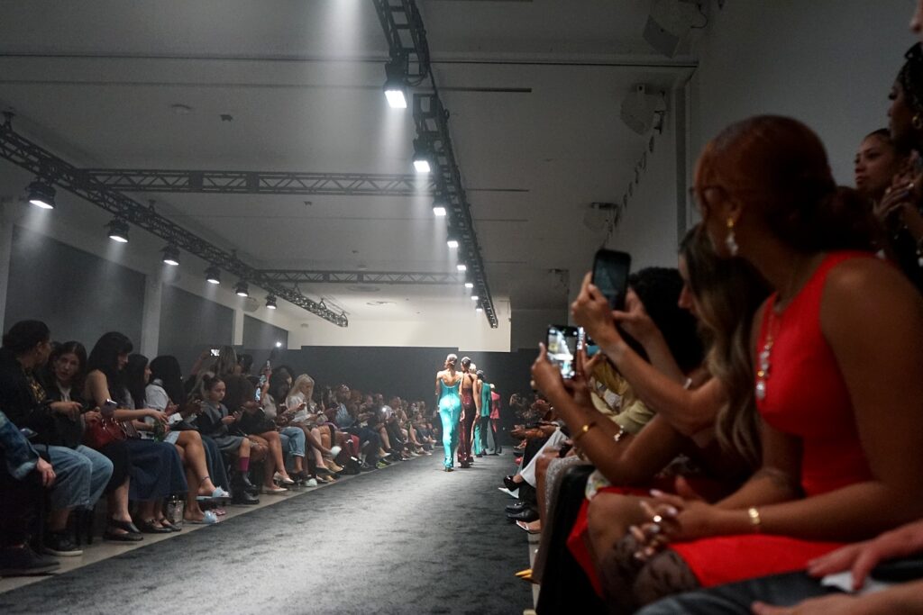 An image of a fashion show where the spotlight is on the runway with models in long dresses walking the catwalk and several people sitting in chairs along the catwalk, many holding their phone and talking photos.