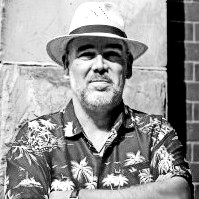 Black-and-white photo of a man with a beard, wearing a patterned shirt and a light-colored fedora. He stands in front of a brick and stone wall with arms crossed, looking at the camera.