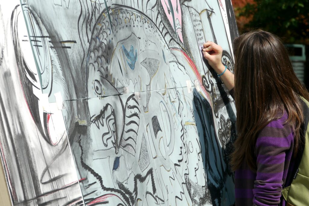 A student with long hair and a striped purple sweater draws on a large outdoor canvas covered in abstract, black-and-white sketch-style illustrations, including a large fish-like figure.