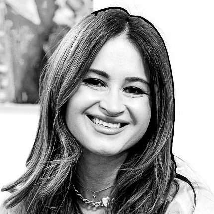 Black-and-white image of a smiling woman with long, wavy hair. She is wearing a sheer, light-colored top and layered necklaces, seated in a brightly lit room with artwork in the background.