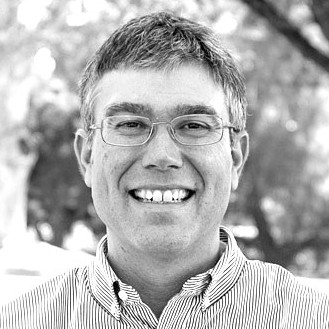 Black-and-white portrait of a smiling man wearing glasses and a striped button-down shirt. He stands outdoors with blurred trees in the background.