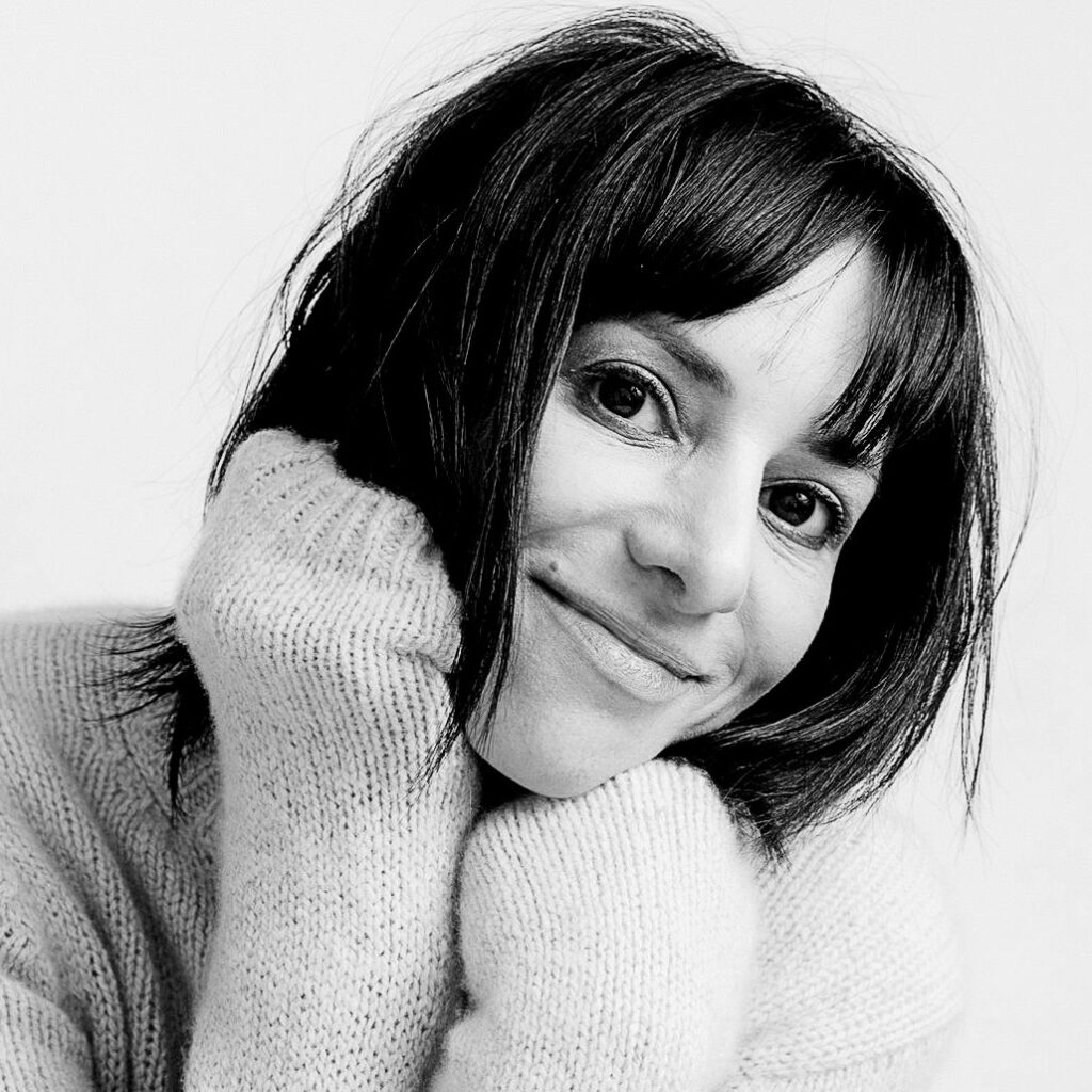 Black-and-white image of a woman with chin-length dark hair and bangs, smiling gently. She is wearing a soft, knit sweater and leaning forward with her hands near her face.