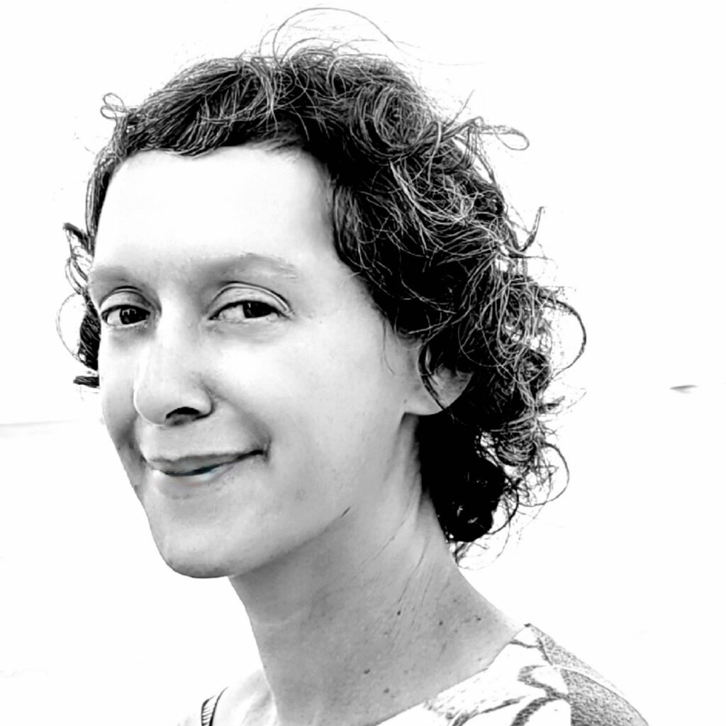 Black-and-white portrait of a woman with short, curly hair, smiling softly. She is wearing a patterned top and standing against a bright, overexposed background.