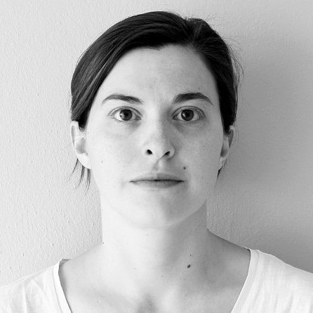 Black-and-white photo of a woman with dark hair pulled back, wearing a plain white shirt. She faces the camera with a neutral expression, standing against a smooth, light-colored wall.