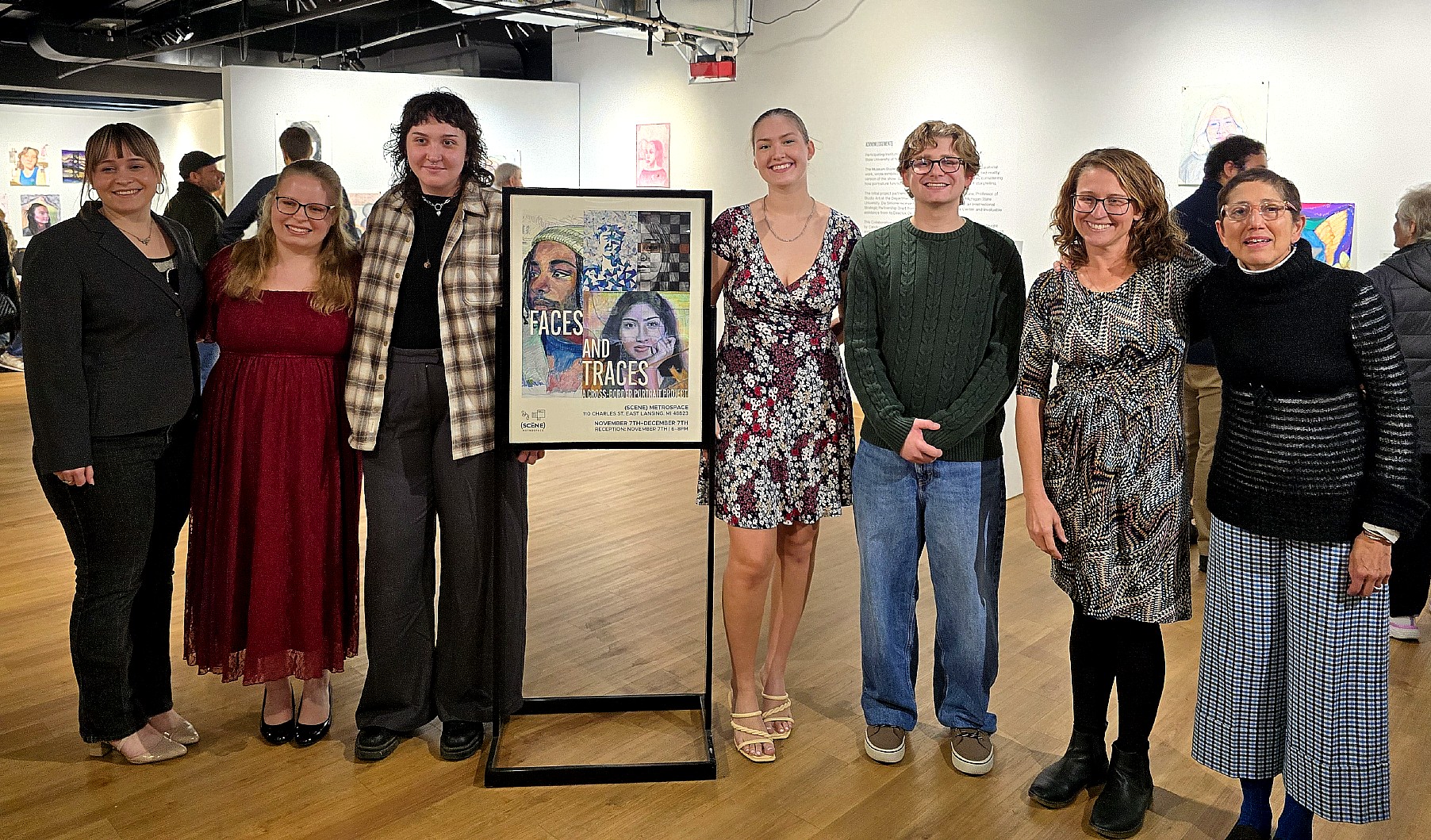 Seven people stand in a row beside the exhibition poster inside the gallery, posing for a group photo with artworks visible behind them.