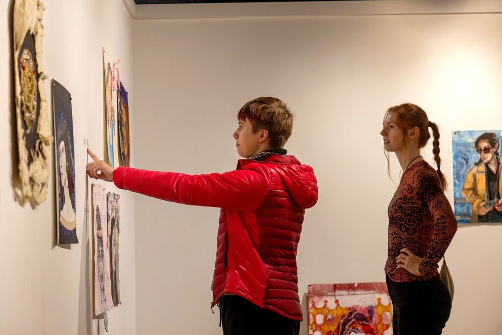 Two people stand in front of a gallery wall; one in a red jacket points toward a label beside a series of portrait artworks while the other watches.