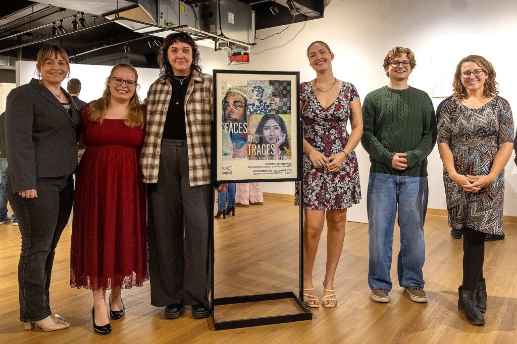Six people stand in a row beside the exhibition poster inside the gallery, posing for a group photo with artworks visible behind them.