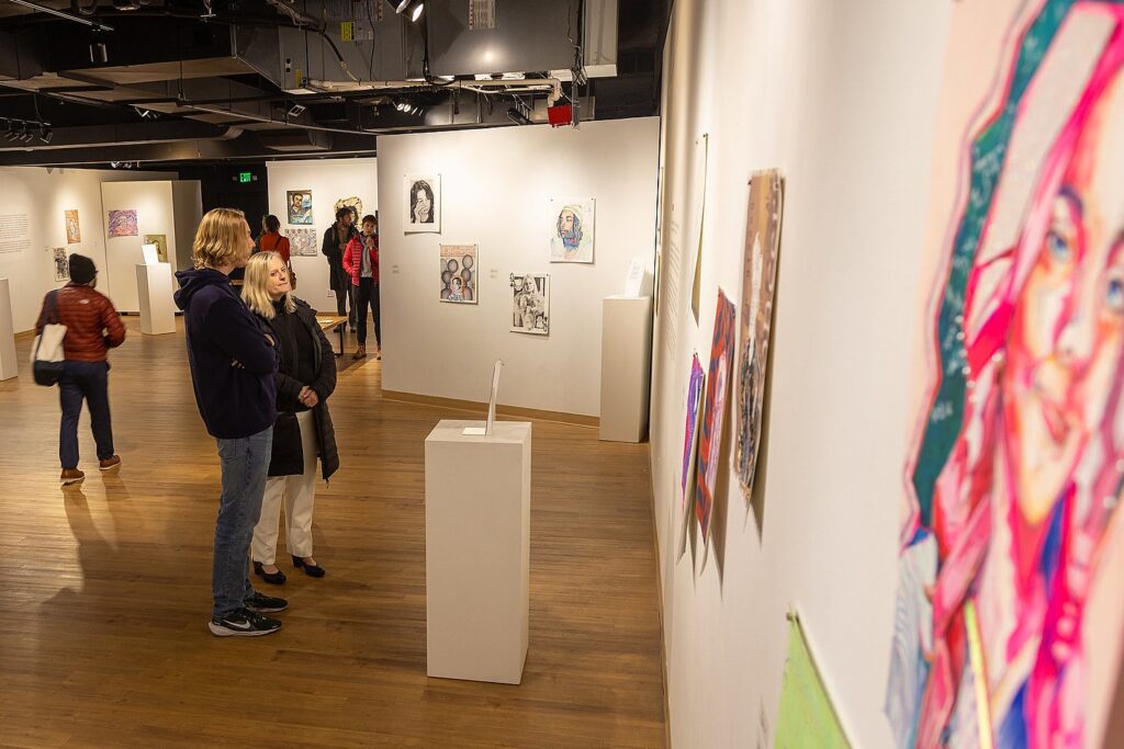 Gallery visitors move through a brightly lit exhibition space, viewing portraits and mixed-media artworks displayed on walls and pedestals.