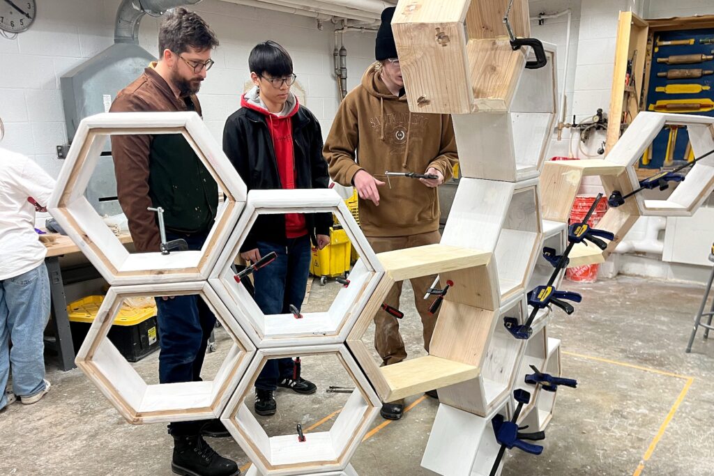 In a workshop, three people stand around a partially assembled wooden sculpture made of large hexagonal frames. Tools, clamps, and wood scraps are scattered throughout the space, and ventilation ducts run across the ceiling.
