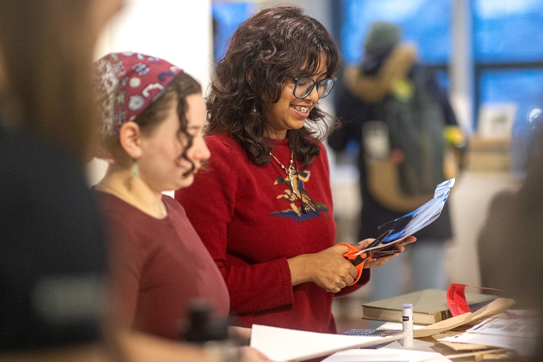 Two participants stand side by side at a worktable, one holding scissors and paper, as they create collage pieces during the Patchwork: Reclaiming Space exhibition.