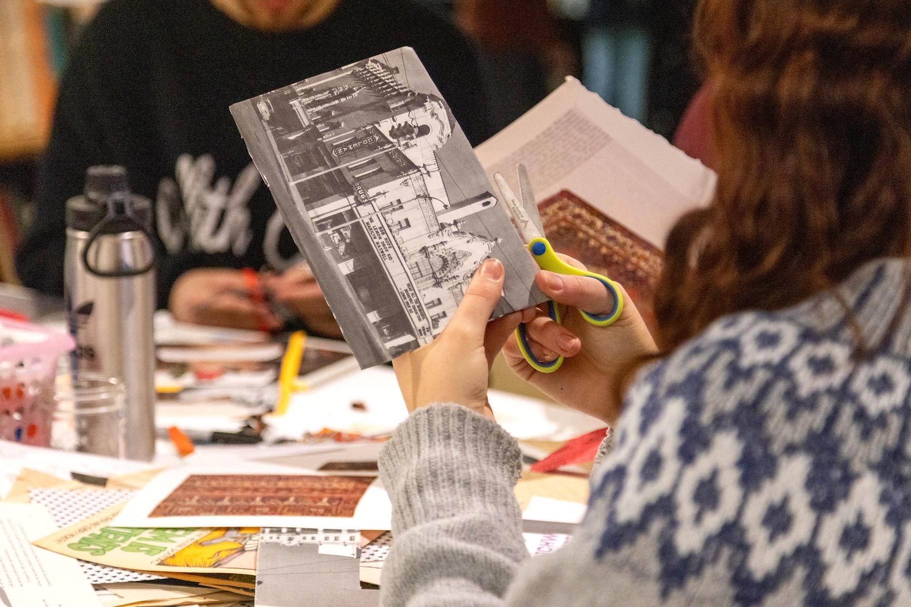 A close-up shows hands cutting a black-and-white printed image with scissors at a table filled with collage materials, books, and paper, highlighting the hands-on, participatory nature of the Patchwork: Reclaiming Space exhibition.