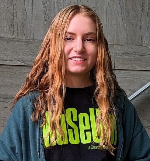 A professional headshot of Rachel Lewis. She has long, wavy light-brown hair and is wearing a dark teal zip-up hoodie over a black t-shirt featuring a lime-green "Museum" logo. She is smiling against a textured grey stone wall background.