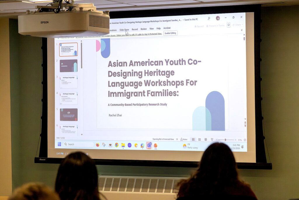 A projected presentation slide titled “Asian American Youth Co-Designing Heritage Language Workshops for Immigrant Families: A Community-Based Participatory Research Study” by Rachel Zhai is displayed at the front of a room, with audience members visible in silhouette.