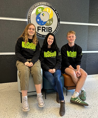 Three people are seated on colorful ottomans in front of the Facility for Rare Isotope Beams (FRIB) logo. They wear matching "Museum" sweatshirts.