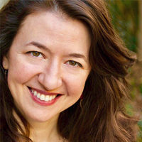 Close-up professional headshot of Alexis Black with long, wavy brown hair, smiling in a natural outdoor setting with soft sunlight.