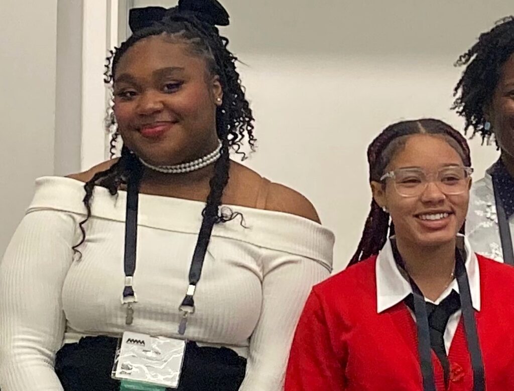 Two students wearing conference lanyards smile at the camera while standing indoors; one wears an off-the-shoulder white top and pearl necklace, and the other wears a red sweater over a white collared shirt.
