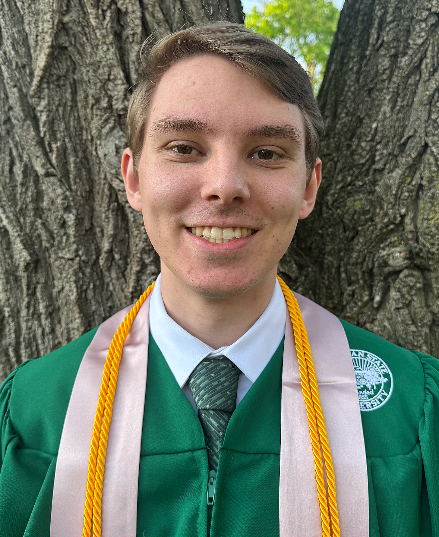Young man in a green graduation gown and tie stands smiling in front of a tree. He wears a pink stole and yellow honors cord, conveying achievement.