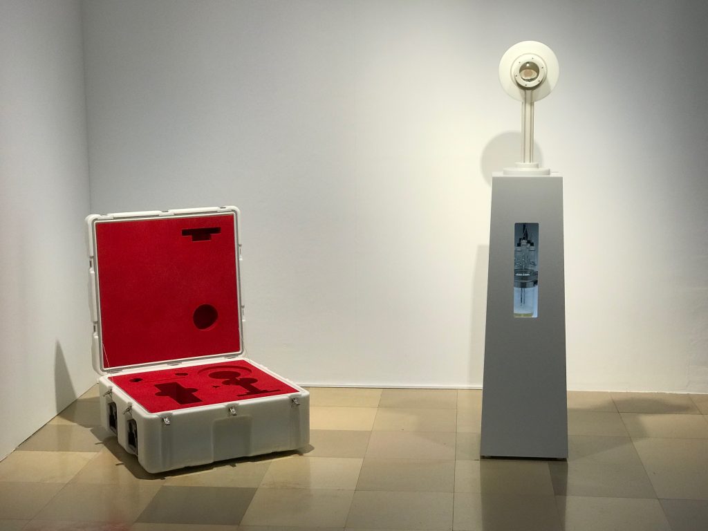 Art exhibit of a white box (left) lined with red padding. Next to it on the right is a white device with a circular part containing a magnified piece of bread in the center of the circle.