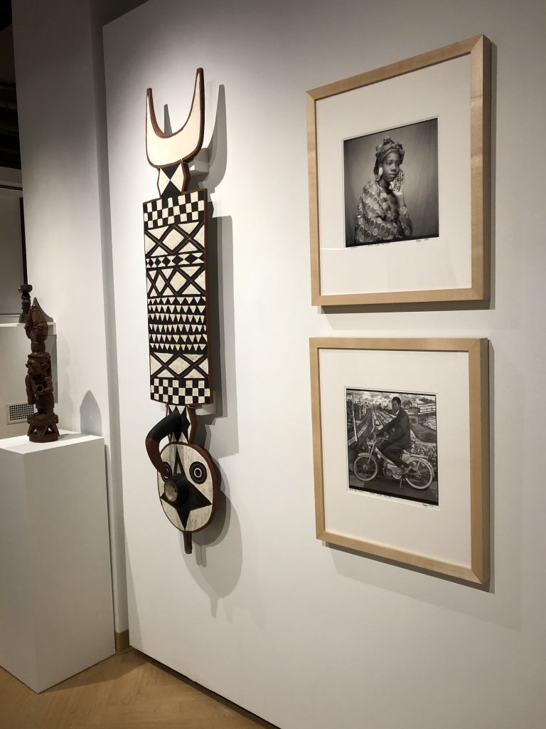 Installation image from "Africa is Not A Country: Artwork from Africa and the African Diaspora from MSU Collections" featuring two black and white portrait photographs, a large wooden mask with a black and white pattern painted on the surface, and a carved wood statue.